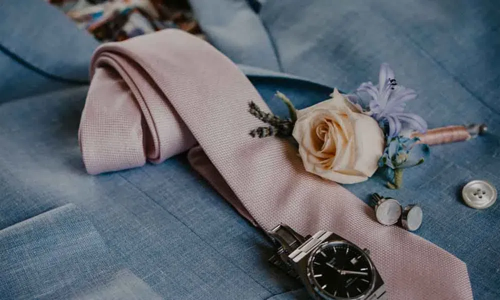 Detailaufnahme eines hellblauen Anzugs mit rosafarbener Krawatte, Uhr, Manschettenknöpfen und einer Ansteckblume aus Rose und Lavendel.