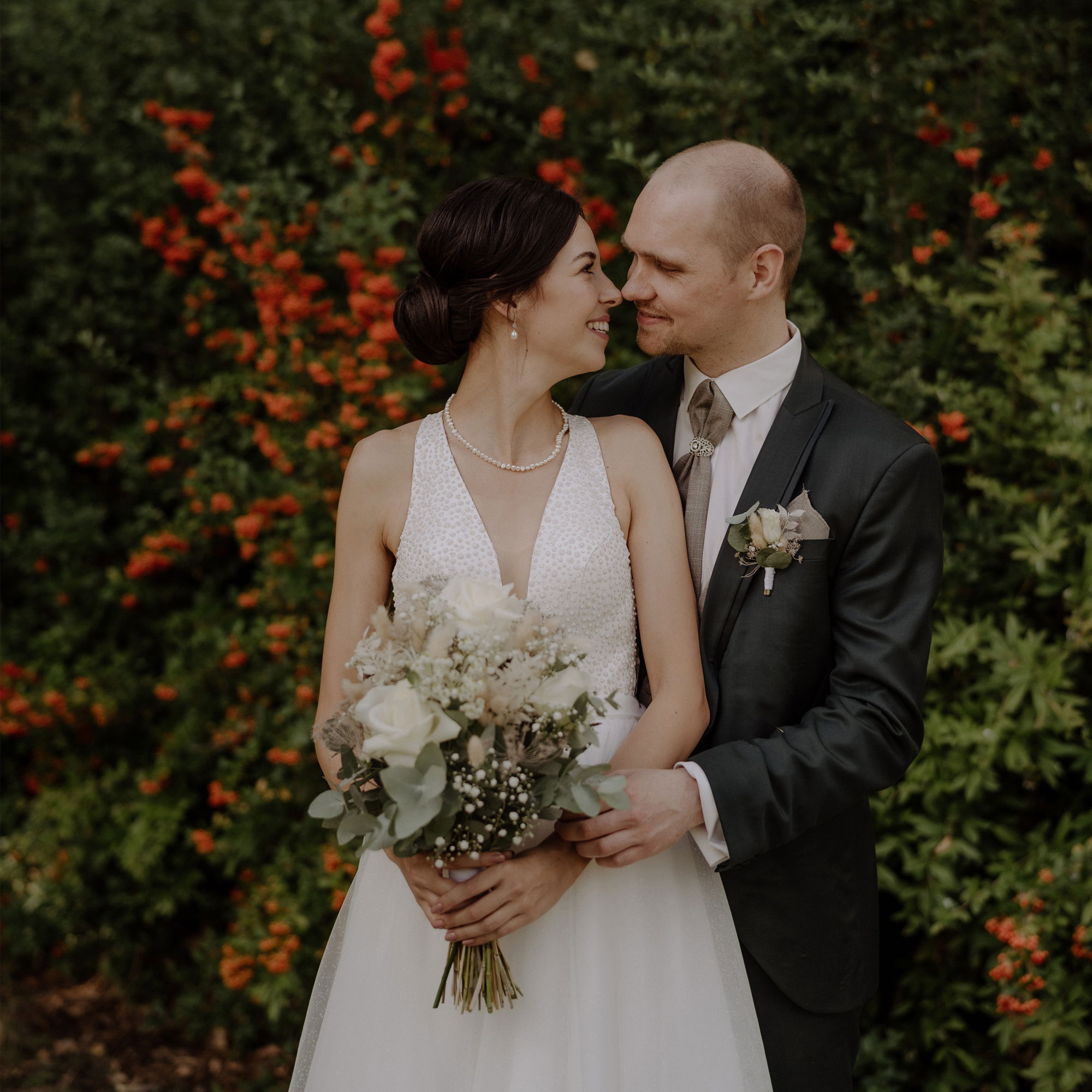 Ein Brautpaar steht eng umarmt vor einer grünen Hecke mit roten Beeren. Die Braut trägt ein elegantes weißes Kleid mit Perlenkette und hält einen Blumenstrauß mit weißen Rosen und Eukalyptus. Der Bräutigam trägt einen dunkelgrünen Anzug mit einer silbernen Krawatte und Boutonniere. Beide lächeln sich liebevoll an.