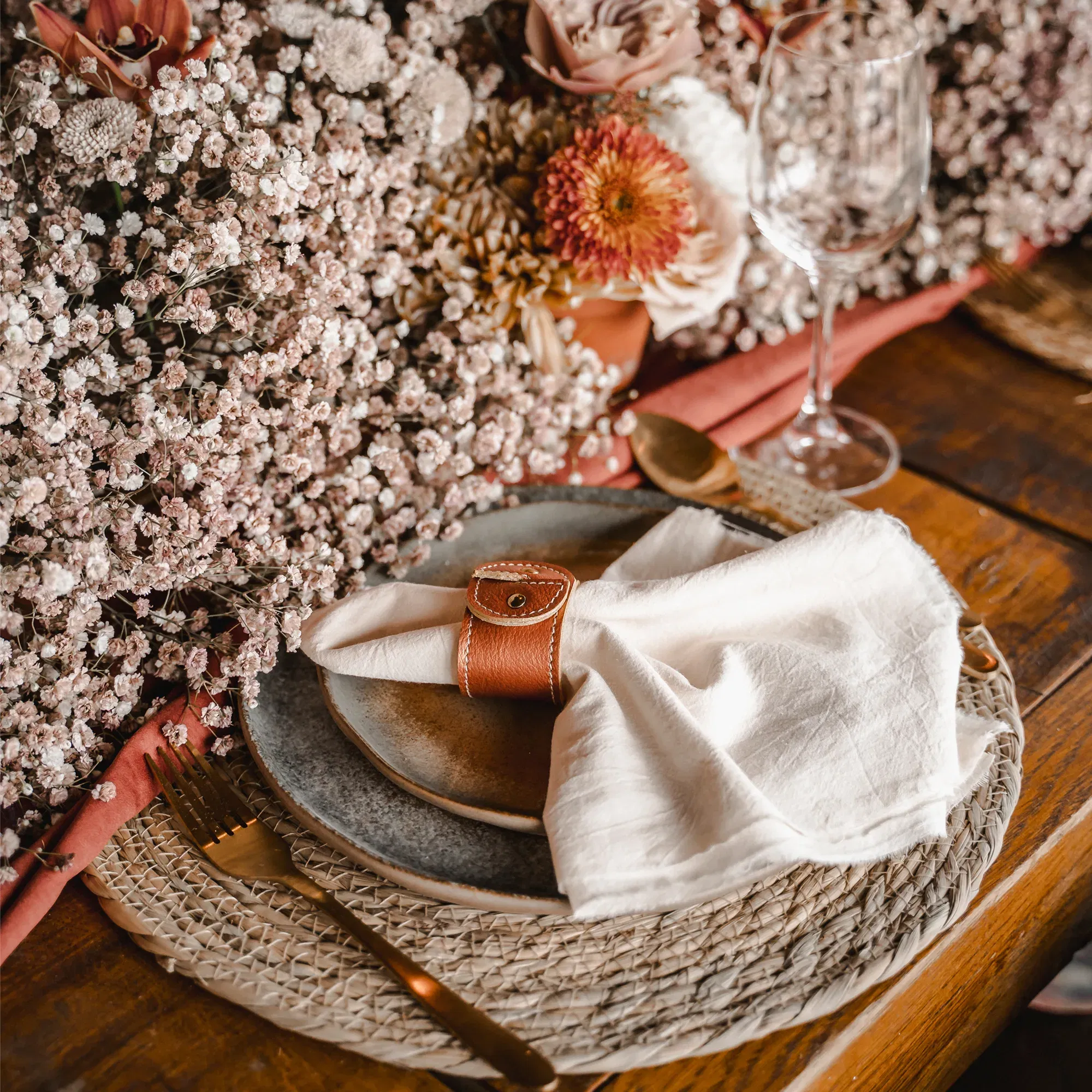 Rustikales Tischgedeck für eine Hochzeit mit einer Stoffserviette in einem braunen Lederring auf einem Keramikteller, arrangiert mit Schleierkraut und Trockenblumen.