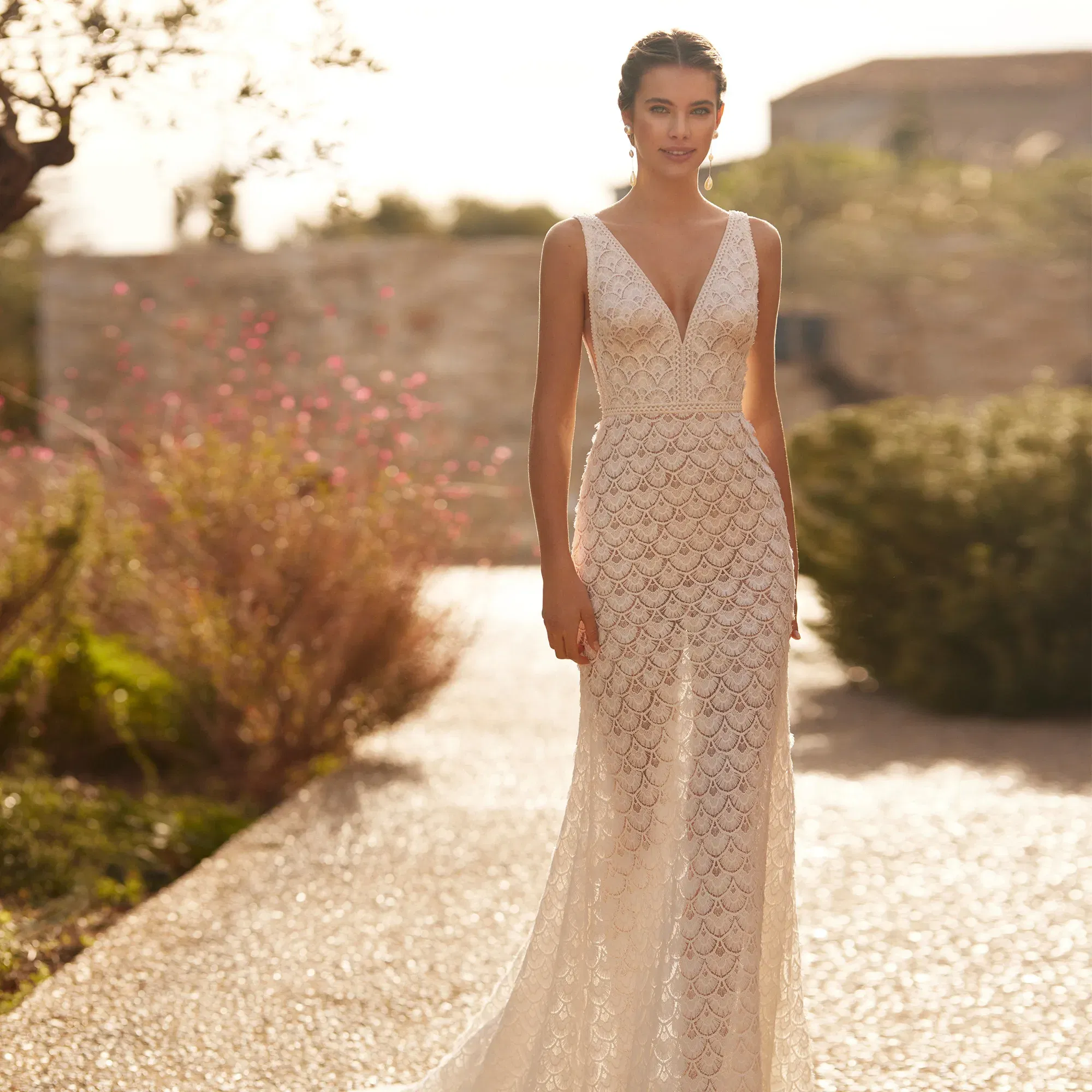 Romantische Braut trägt ein ärmelloses Spitzenkleid mit tiefem V-Ausschnitt, posierend in einem sonnendurchfluteten Garten.