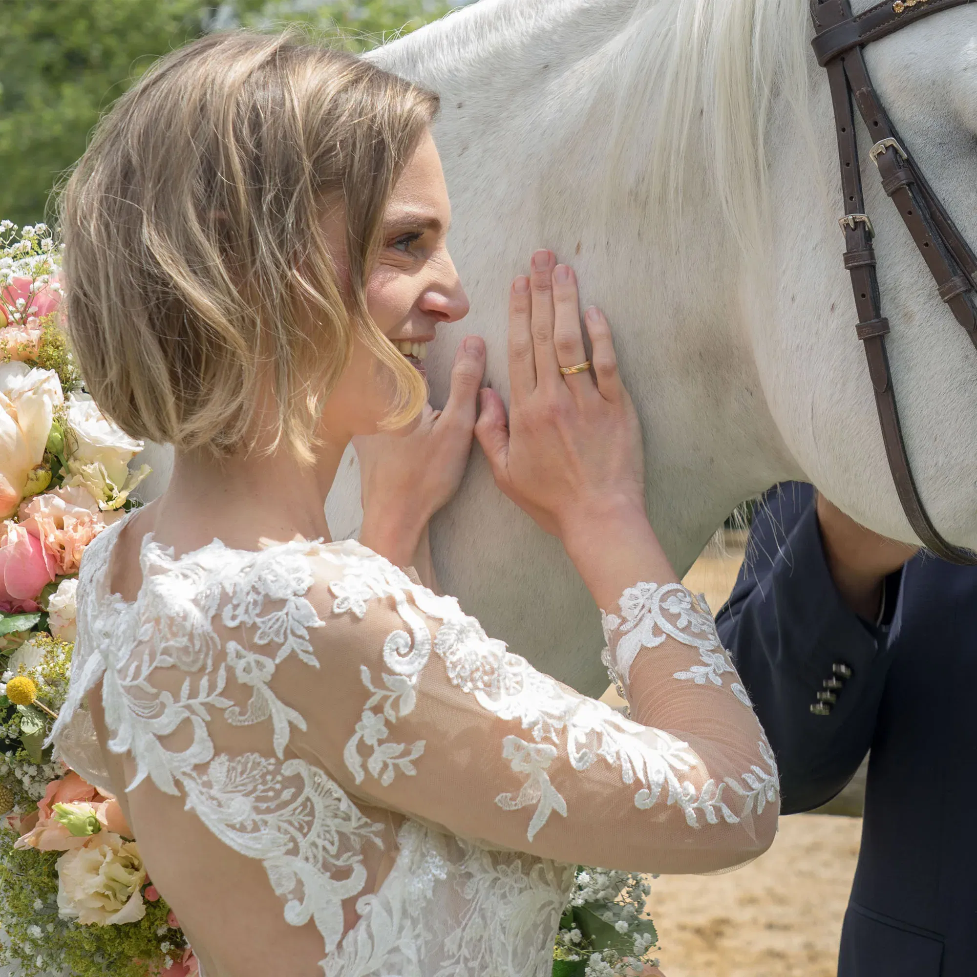 Eine Braut in einem eleganten Spitzenkleid lehnt sich lächelnd gegen den Hals eines weißen Pferdes und streichelt es sanft. Das Pferd trägt einen Blumenkranz, und die Szene spielt sich draußen in einer grünen Umgebung ab.