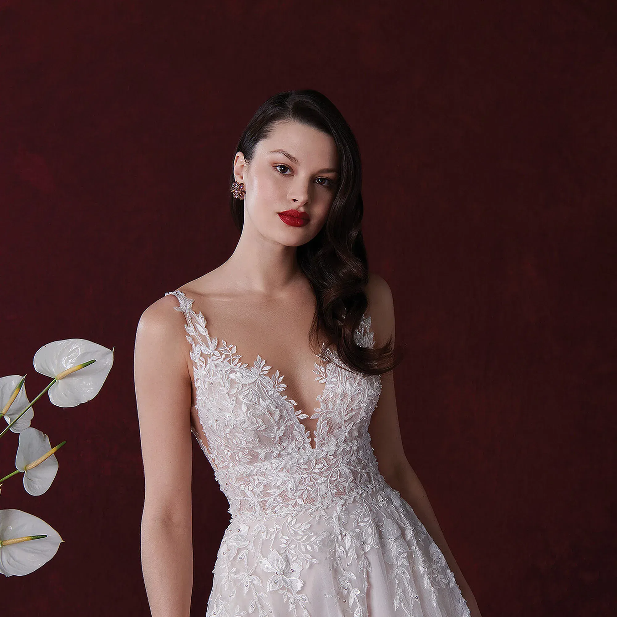 Braut in einem kunstvoll bestickten Spitzenkleid von Justin Alexander mit tiefem V-Ausschnitt, steht vor einem dunkelroten Hintergrund. Sie trägt leuchtend roten Lippenstift und hat ihr Haar in glamourösen Wellen über eine Schulter gelegt.
