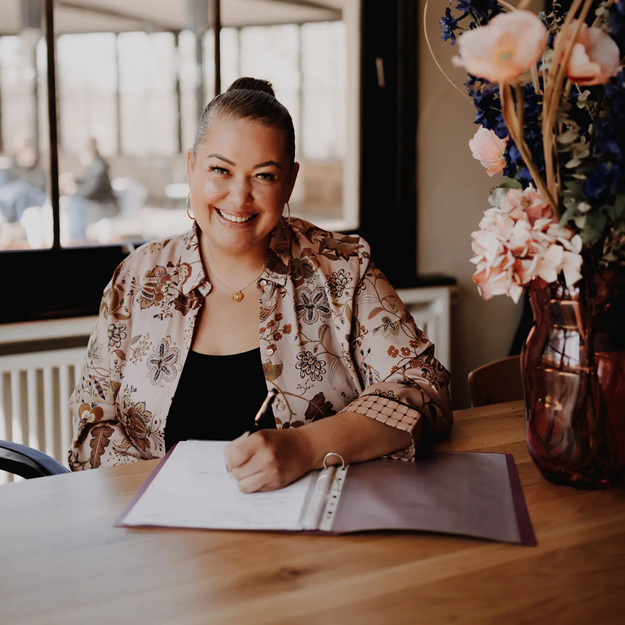 Eine Frau mit freundlichem Lächeln sitzt an einem Tisch, schreibt in einen Ordner und trägt eine Bluse mit Blumenmuster. Auf dem Tisch steht eine große Vase mit rosa und blauen Blumen.