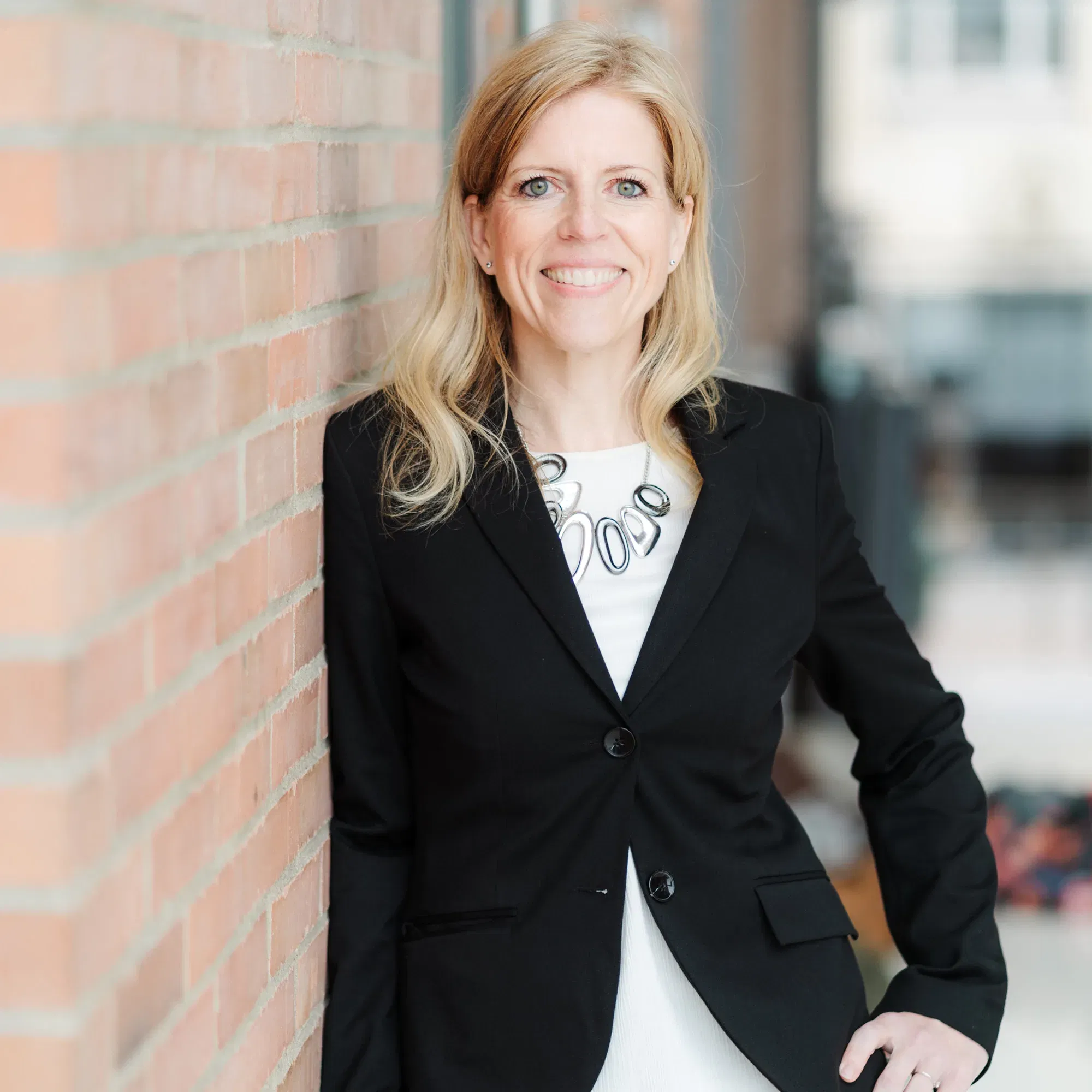 Porträt einer Frau mit langem, blonden Haar, die in einem schwarzen Blazer an einer Backsteinwand lehnt. Sie lächelt freundlich und trägt eine moderne Halskette und weiße Kleidung unter dem Blazer.