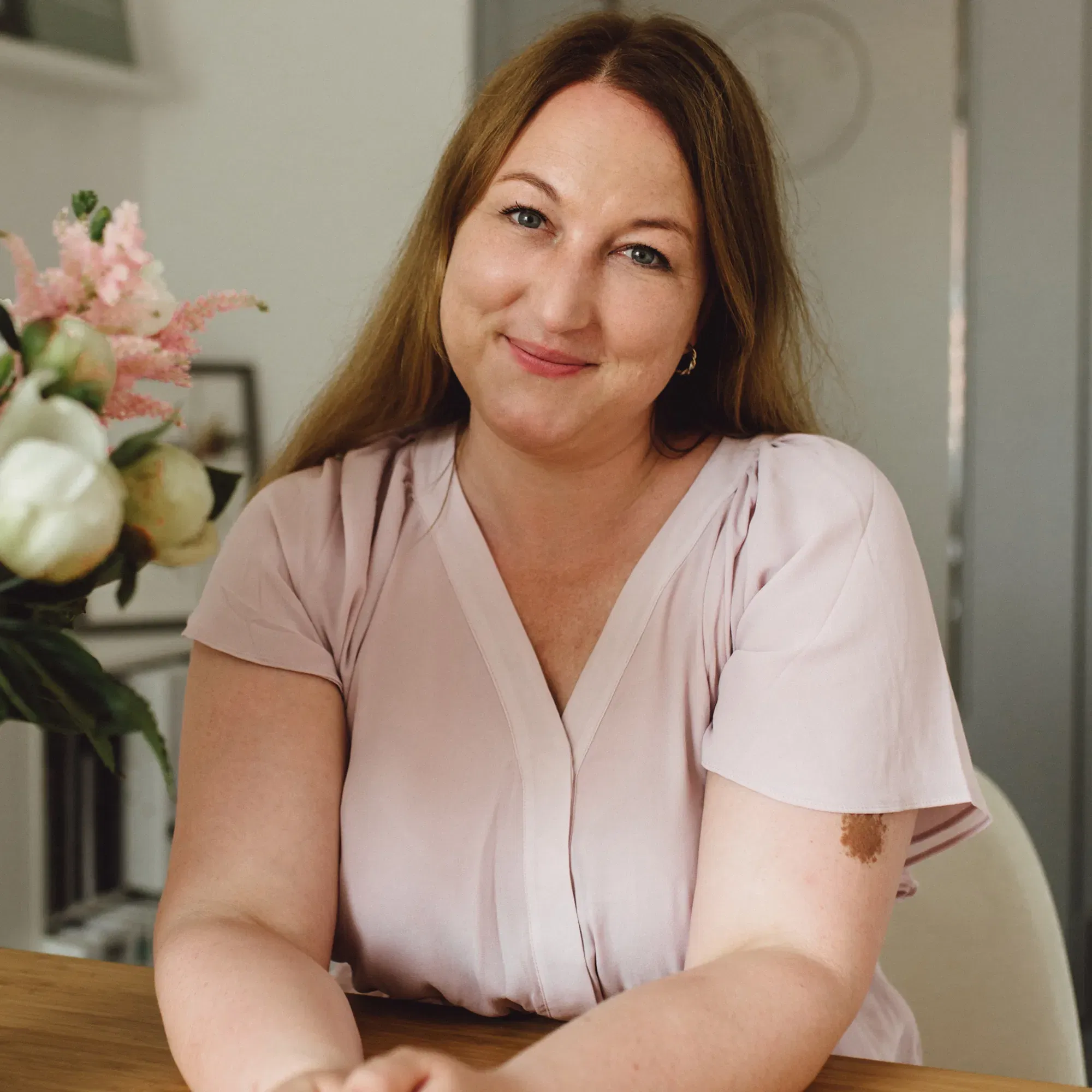 Porträt einer Frau mit langen, braunen Haaren, die in einem hellrosa Bluse an einem Tisch sitzt. Sie lächelt sanft in die Kamera, während eine Blumenvase neben ihr auf dem Tisch steht.