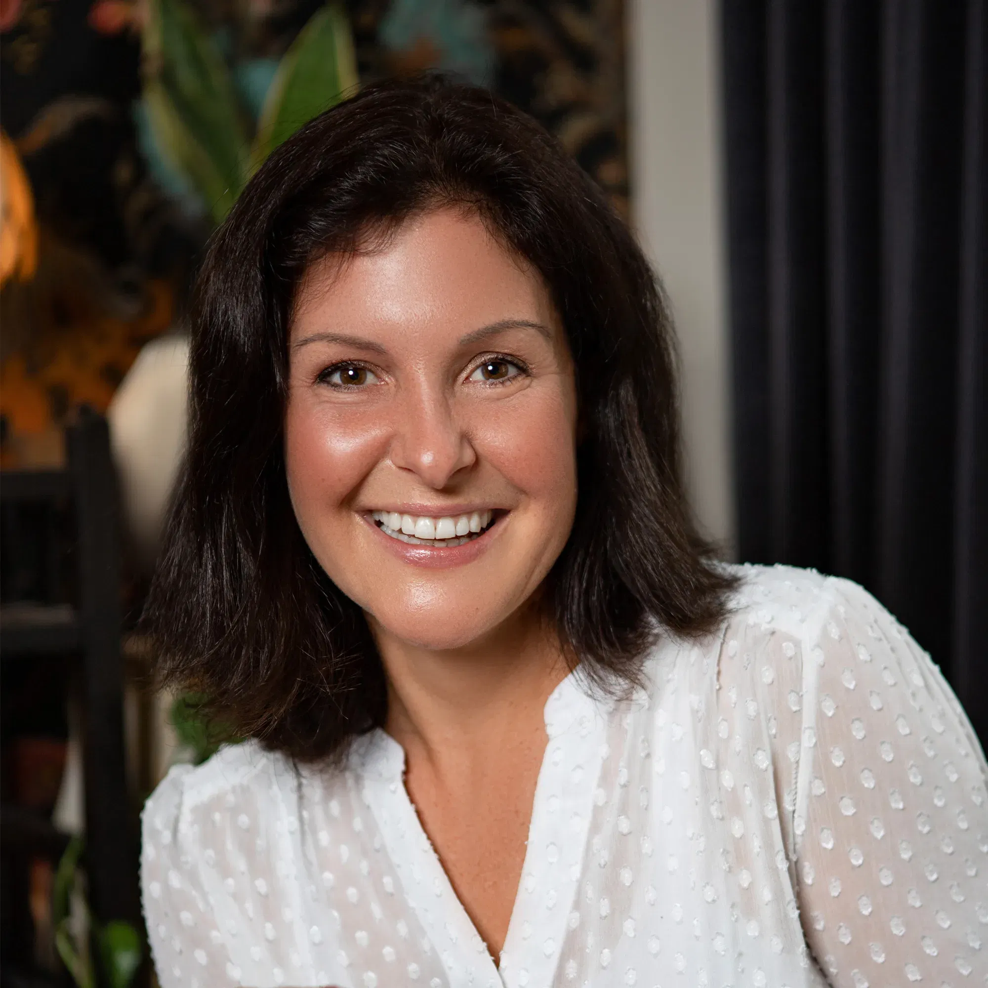 Porträt einer lächelnden Frau mit dunklem, schulterlangem Haar in einer weißen Bluse mit dezentem Punktmuster. Der Hintergrund ist stilvoll mit Pflanzen dekoriert.