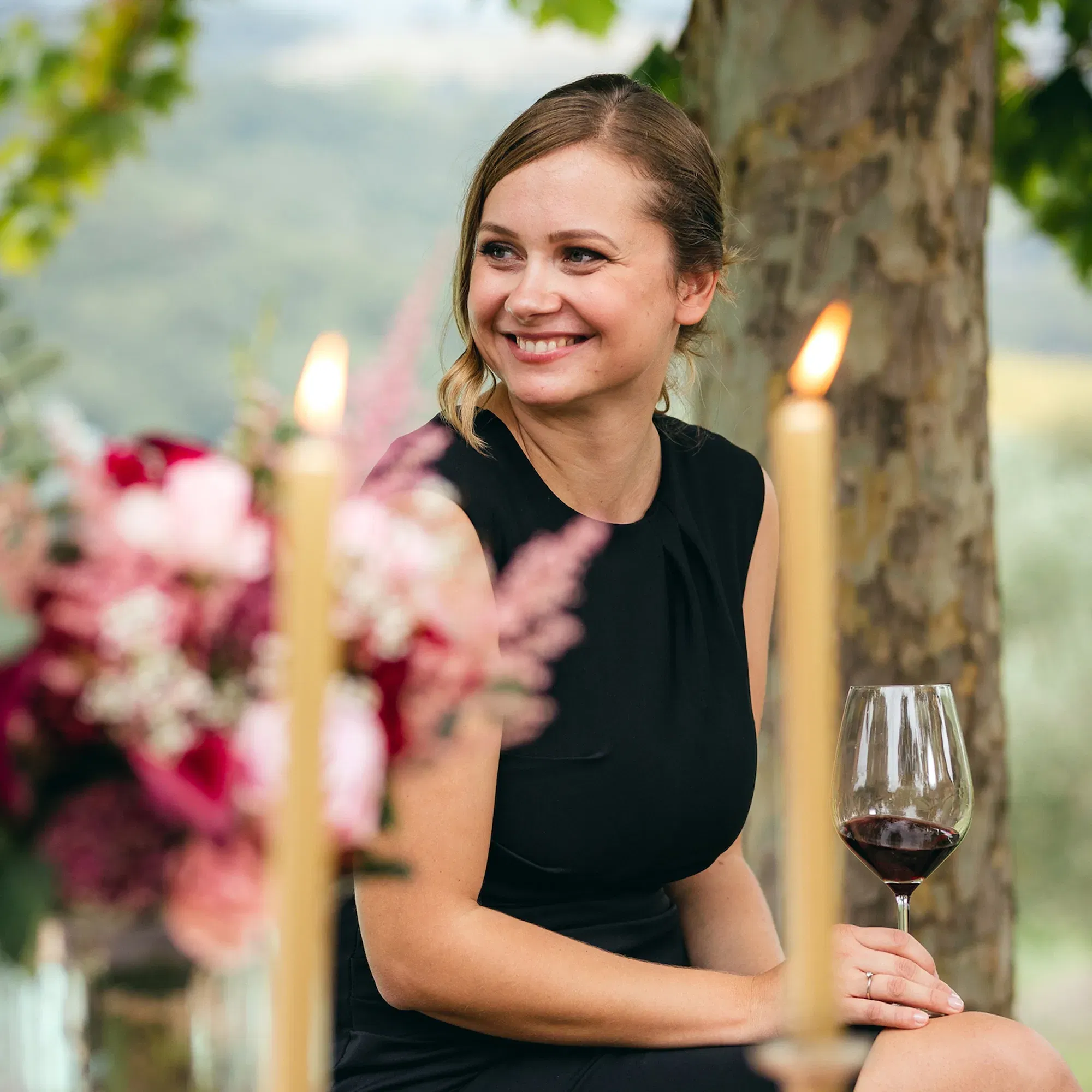 Porträt einer Frau in einem eleganten schwarzen Kleid, die lächelnd an einem Tisch mit blühenden Blumen und brennenden Kerzen sitzt. Sie genießt ein Glas Rotwein in einer idyllischen Outdoor-Umgebung.