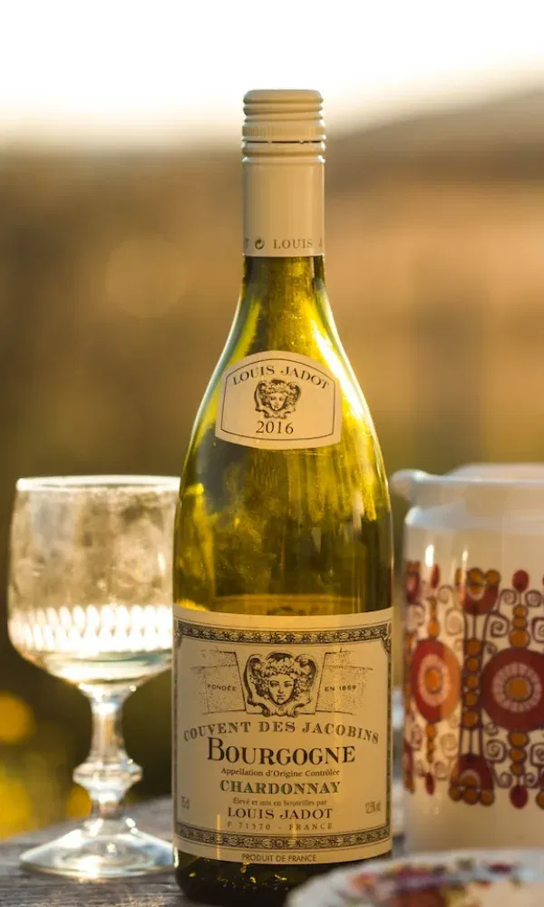 Ein Glas Weißwein, ein bunter Krug und eine Flasche französischer Chardonnay mit Etikett stehen auf einem gedeckten Tisch im Freien.