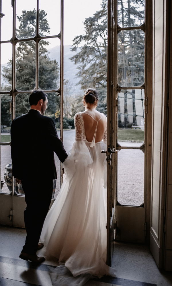 Das Brautpaar geht durch hohe Glastüren in den Garten hinaus, die Braut in einem fließenden Brautkleid, der Bräutigam in einem eleganten schwarzen Anzug.