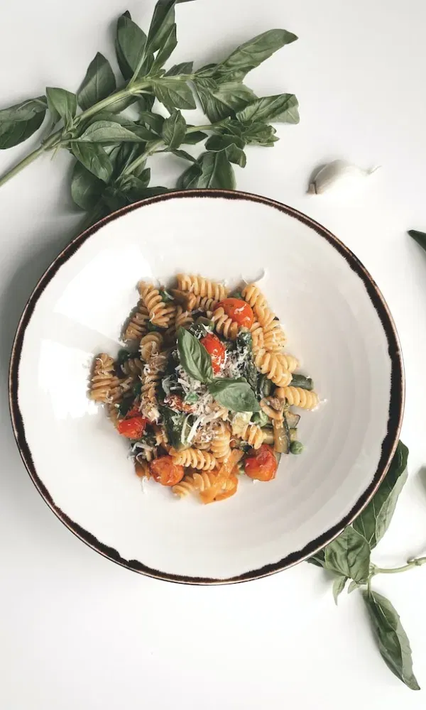 Vegane Fusilli mit Zucchini, Tomaten, Erbsen und geriebenem Käse, angerichtet in einem tiefen Teller und umgeben von frischem Basilikum.