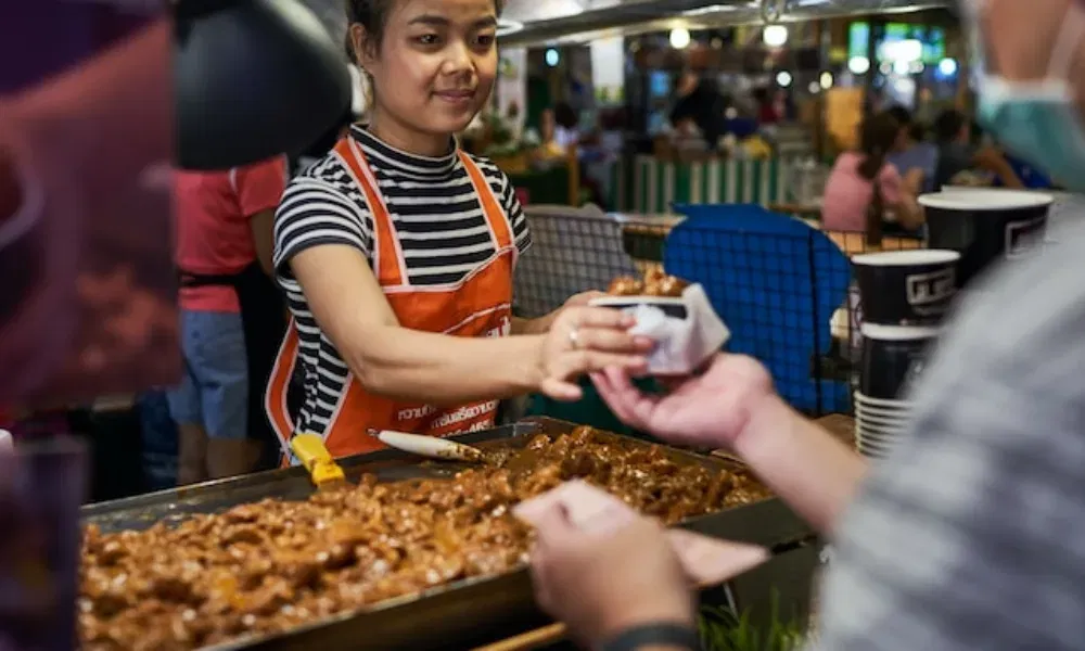 Junge Frau mit gestreiftem Shirt und roter Schürze serviert warmes Essen an einem Street-Food-Stand bei Nachtmarkt-Atmosphäre.