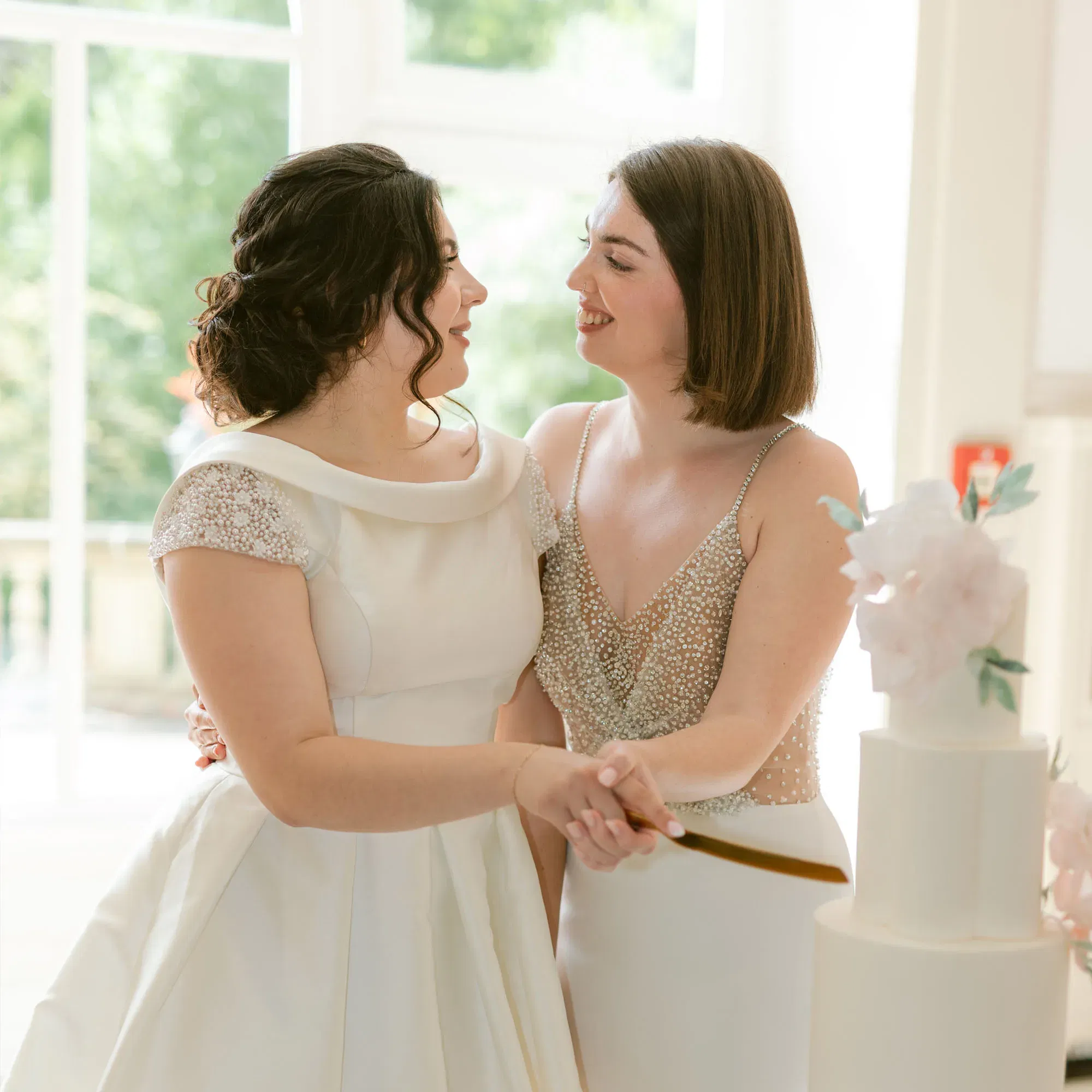 Ein emotionaler Moment zwischen zwei Bräuten, die sich anlächeln, während sie Hand in Hand ihre Hochzeitstorte anschneiden. Beide tragen stilvolle Brautkleider, eines mit Perlstickereien und das andere mit einem eleganten, minimalistischen Schnitt. Sonnenlicht durchflutet den Raum durch die großen Fenster im Hintergrund.