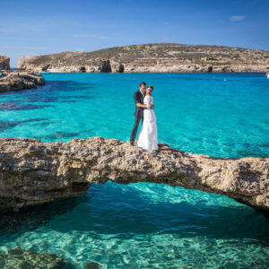 Ein Brautpaar steht auf einem schmalen Felsvorsprung über dem klaren, türkisblauen Wasser der Blue Lagoon auf der Insel Comino, Malta. Die Braut trägt ein weißes Hochzeitskleid, der Bräutigam einen dunklen Anzug. Im Hintergrund erstreckt sich die felsige Landschaft unter einem strahlend blauen Himmel.