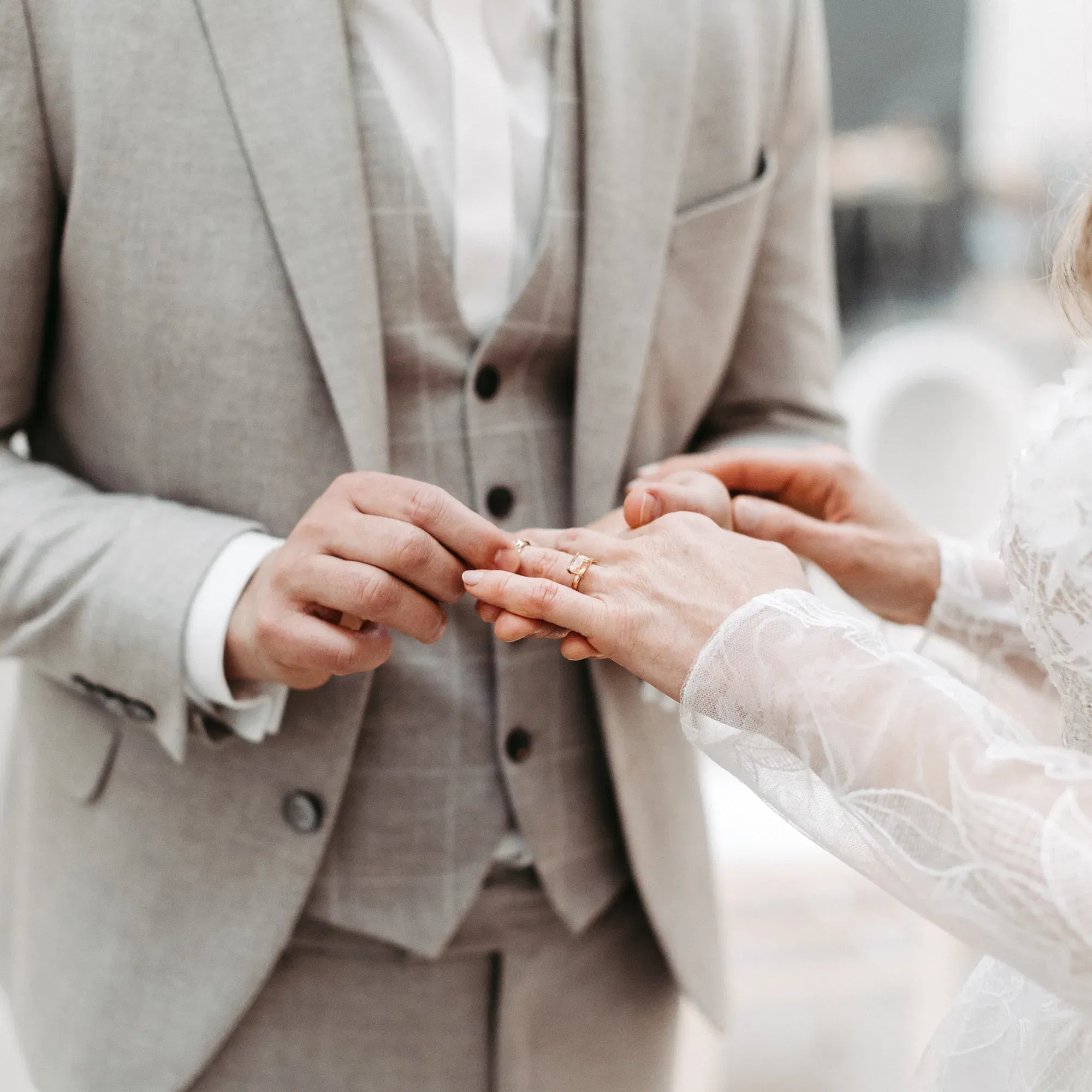 Ein Bräutigam in einem eleganten, hellgrauen Anzug schiebt seiner Braut den Ehering über den Finger. Die Braut ist in einem weißen Spitzenkleid gekleidet, während das Bild den intimen Augenblick der Trauung einfängt.