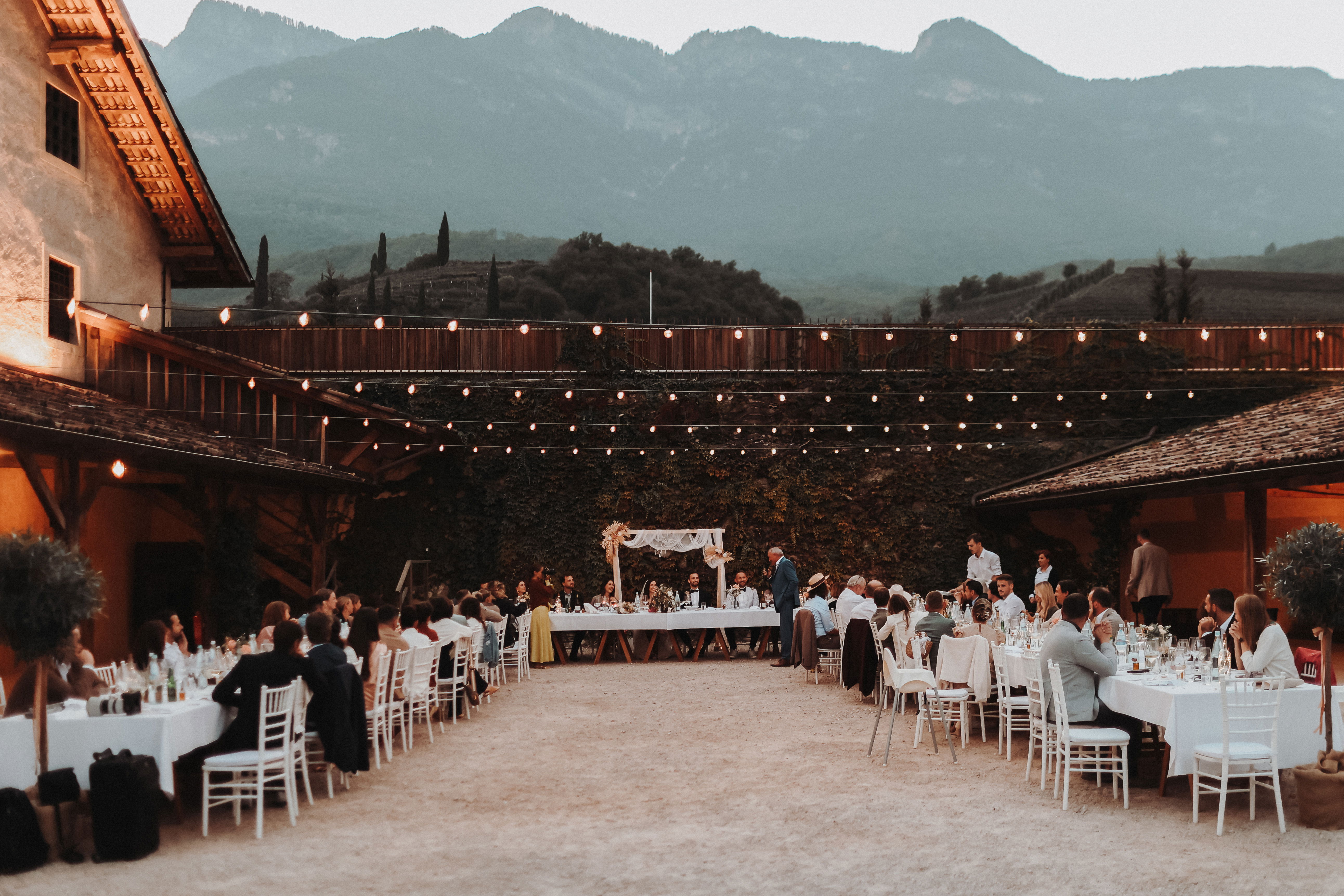Hochzeitsempfang im Freien mit Tischen in U-Form, dekoriert mit weißen Tischdecken und Stühlen. Beleuchtet von Lichterketten, vor einer malerischen Bergkulisse, die eine entspannte, festliche Atmosphäre verleiht.