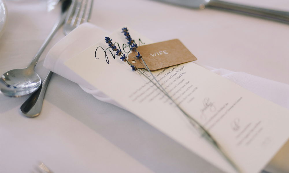 Detailaufnahme einer Hochzeitstafel mit einem Menü auf heller Serviette. Eine braune Namenskarte mit der Aufschrift "WIFE" und ein Lavendelzweig sind darauf dekorativ befestigt. Die stilvolle Tischdeko wird von glänzendem Besteck und einer weißen Tischdecke abgerundet.