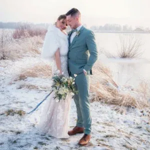 Winterliches Brautpaar am Rand eines gefrorenen Sees. Die Braut in einem spitzenbesetzten Kleid und einer pelzigen Stola lehnt sich lächelnd an den Bräutigam, der einen blauen Anzug trägt. Ihr Blumenstrauß mit weißen Rosen und grünen Akzenten spiegelt den frostigen Charme der Umgebung wider.