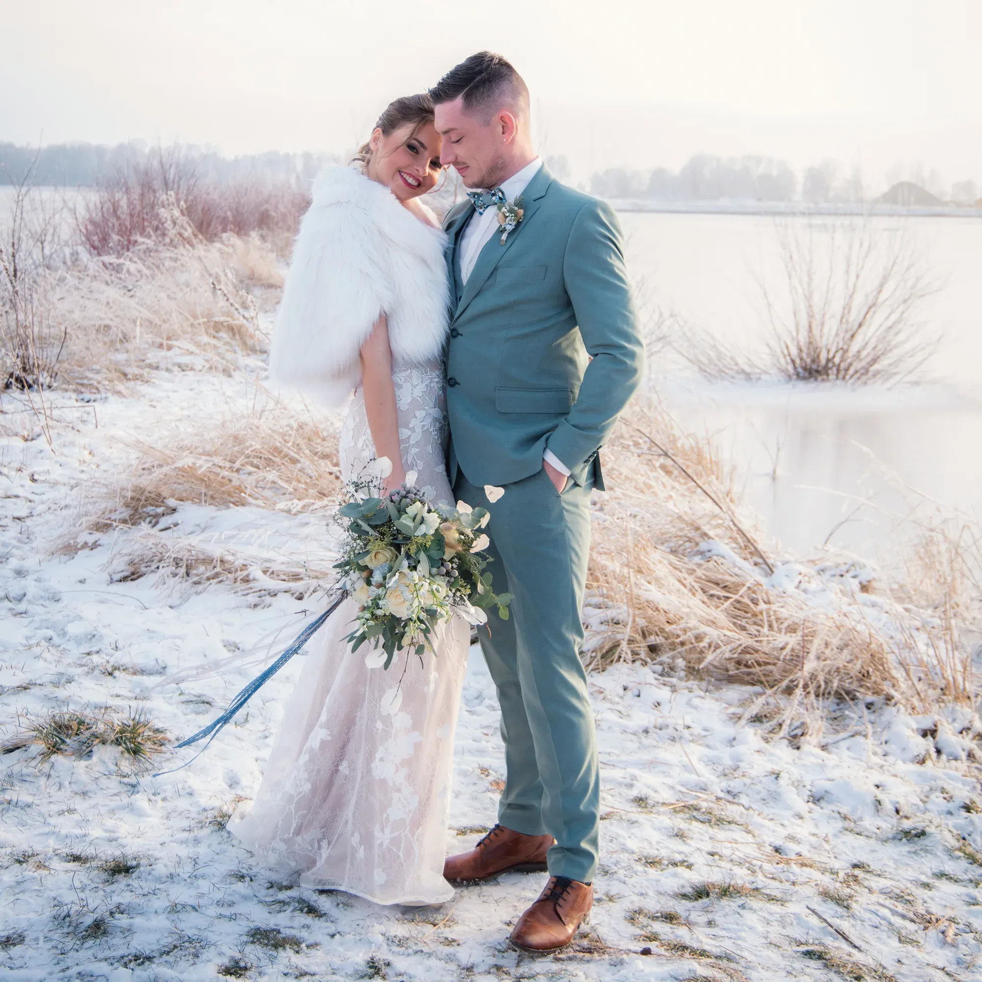 Winterliches Brautpaar am Rand eines gefrorenen Sees. Die Braut in einem spitzenbesetzten Kleid und einer pelzigen Stola lehnt sich lächelnd an den Bräutigam, der einen blauen Anzug trägt. Ihr Blumenstrauß mit weißen Rosen und grünen Akzenten spiegelt den frostigen Charme der Umgebung wider.