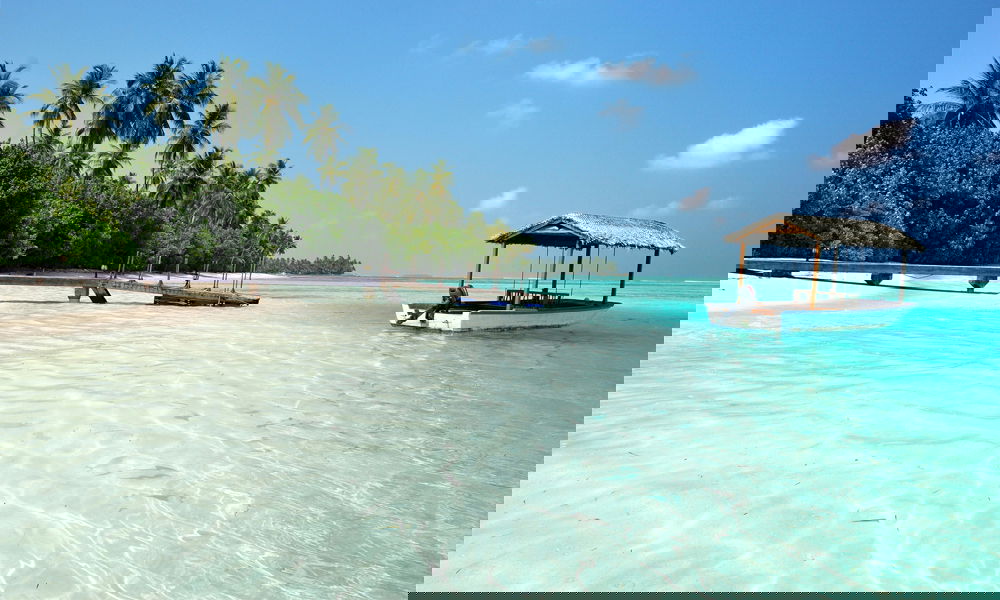 In einer tropischen Lagune liegt ein überdachtes Boot vor Anker, während ein Steg vom palmengesäumten Ufer ins flache Wasser ragt.