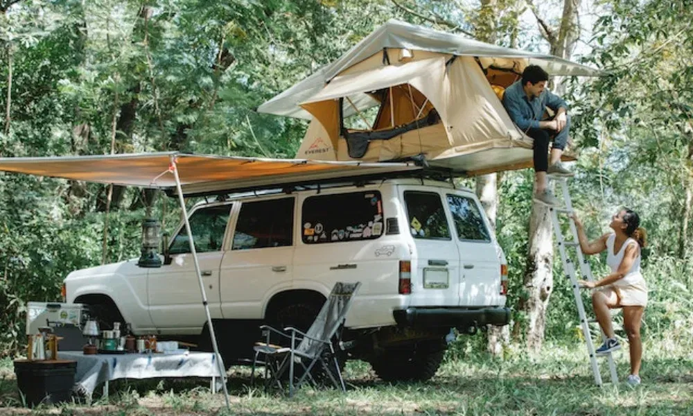 Ein Mann sitzt im Dachzelt auf einem SUV, während eine Frau lachend die Leiter hinaufsteigt – umgeben von Wald.