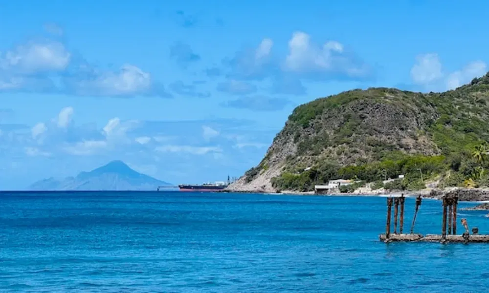 Karibische Meereslandschaft mit einem bewachsenen Hügel, Schiffsanleger, ruhigem Wasser und einer fernen Insel unter blauem Himmel.