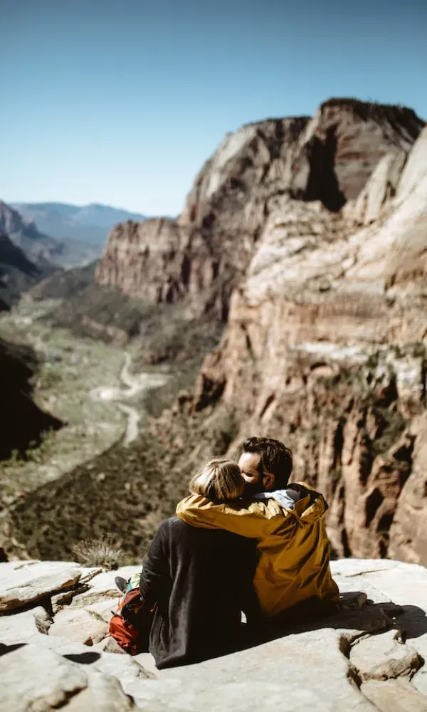 Zwei Menschen genießen eine innige Umarmung auf einer Klippe mit spektakulärer Aussicht über ein Gebirgstal in wilder Natur.