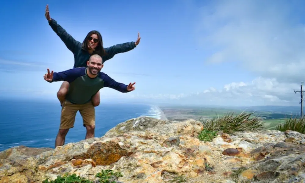 Fröhliches Paar auf einem felsigen Aussichtspunkt mit Blick aufs Meer: Der Mann trägt die Frau huckepack, beide genießen den Moment.