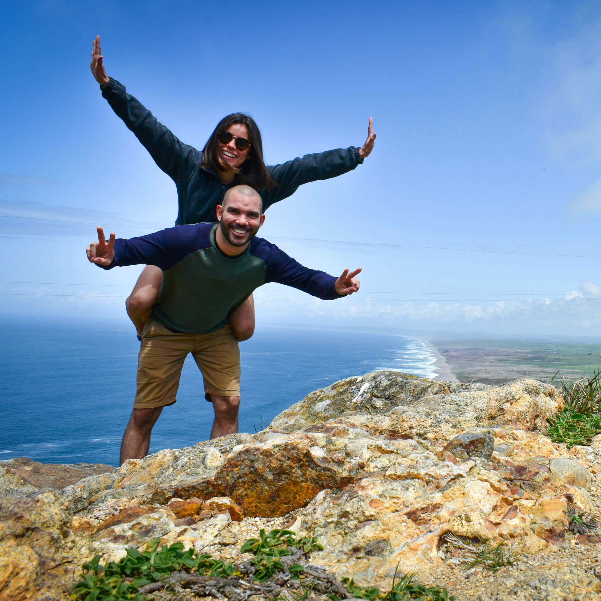 Glückliches Paar beim Wandern: Der Mann trägt seine Partnerin auf den Schultern, beide zeigen fröhlich in die Kamera mit Meer und Küste im Hintergrund.