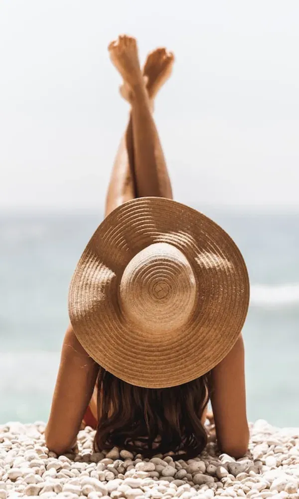 Entspannter Strandmoment: Eine Frau mit langen Haaren ruht auf runden Steinen, der Blick geht aufs Wasser, das Gesicht vom Hut bedeckt.