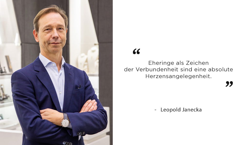 Leopold Janecka, ein Juwelier im Anzug, steht in einem modernen Geschäft und spricht über die emotionale Bedeutung von Eheringen.