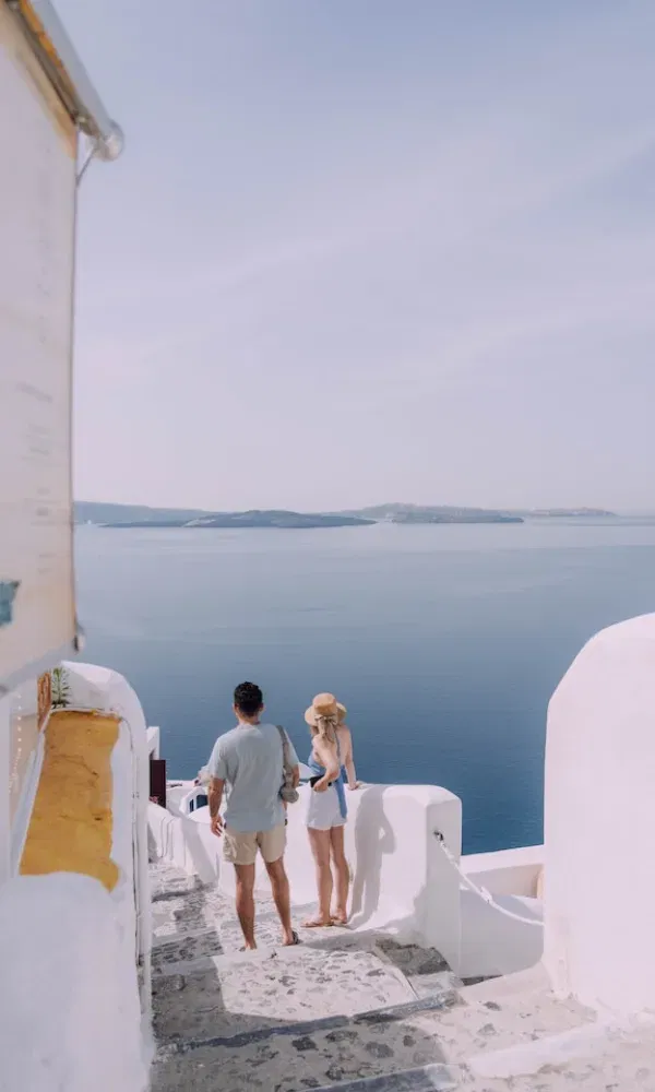 Zwei Menschen genießen den Ausblick über das Ägäische Meer, während sie durch die typischen Gassen von Santorini mit weißen Wänden und Naturstein laufen.