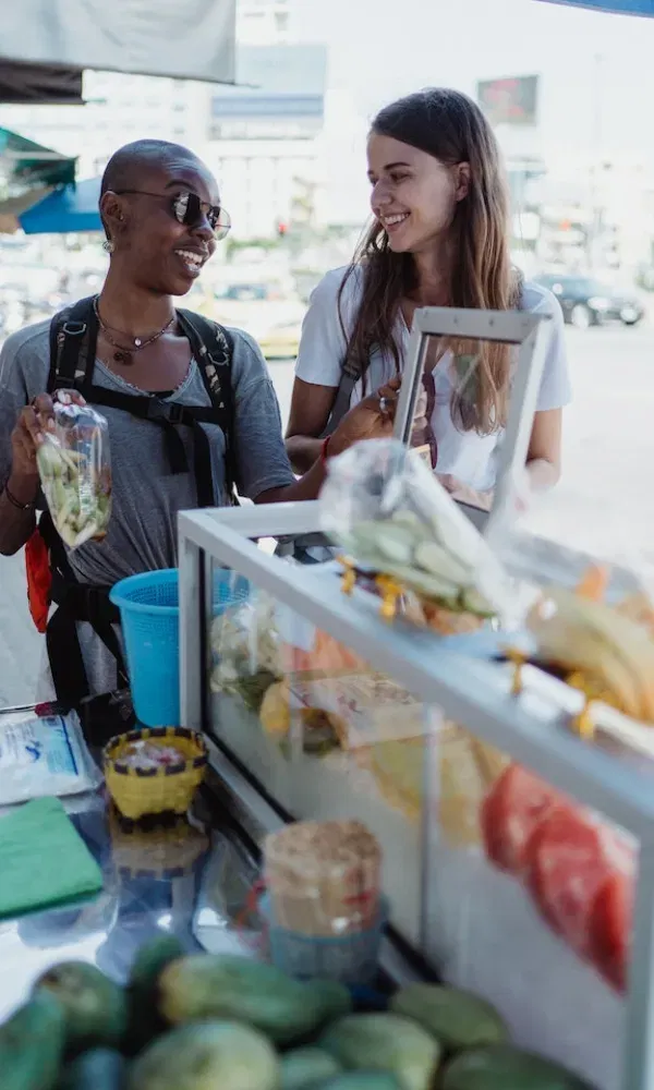 Reiseszene in der Stadt: Zwei Freundinnen stehen an einem Obststand, lachen gemeinsam und zeigen auf tropische Früchte in der Auslage.