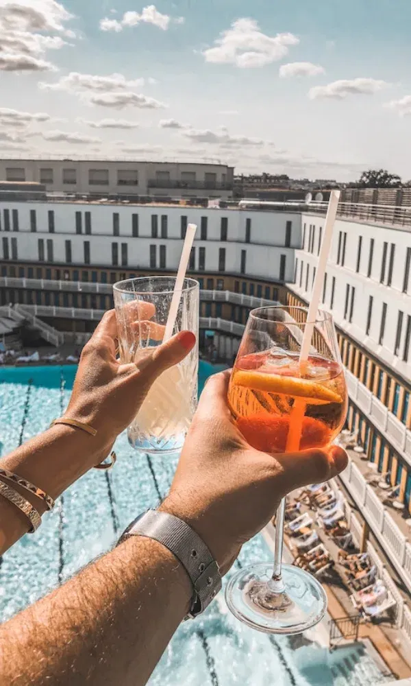 Aussicht von oben auf ein Schwimmbad, während zwei Hände kühle Sommerdrinks mit Strohhalm in die Luft halten.
