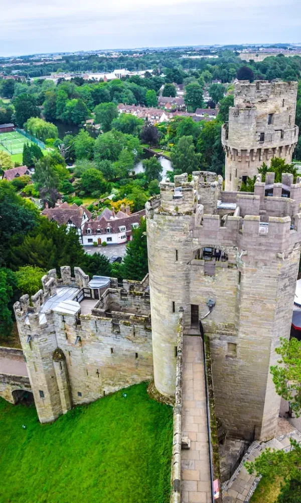 Blick vom Turm einer mittelalterlichen Burg auf ein englisches Dorf mit vielen Bäumen und gepflegtem Rasen – vermutlich Warwick Castle.