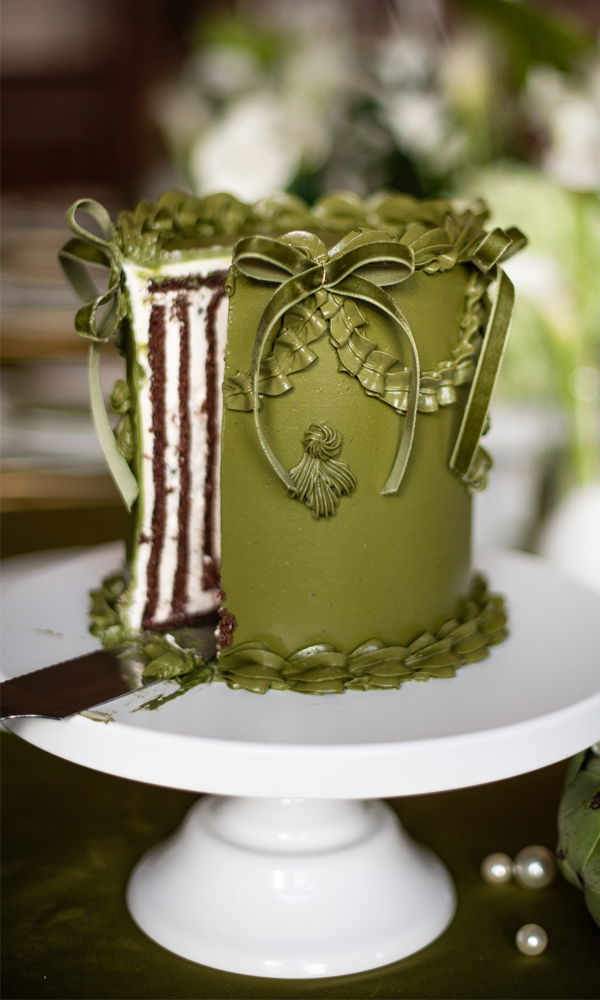 Opulente, einlagige Hochzeitstorte in tiefem Olivgrün mit filigranen Fondant-Verzierungen, darunter Schleifen, geflochtene Bänder und kleine Tasseln. Ein angeschnittener Bereich offenbart eine gestreifte Schokoladen-Vanille-Füllung. Die Szenerie ist in einem edlen, grünen Farbkonzept gehalten.