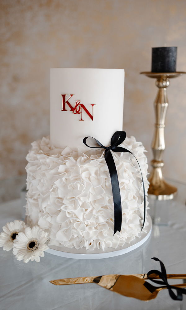Edle Hochzeitstorte mit zwei Ebenen: Die untere Etage ist mit filigranen, weißen Zuckerrüschen bedeckt, während die obere glatt bleibt und mit den roten Initialen „K & N“ verziert ist. Eine schwarze Satinschleife setzt einen stilvollen Akzent. Die Torte ist auf einem schlichten weißen Tablett platziert, umgeben von weißen Blumen.