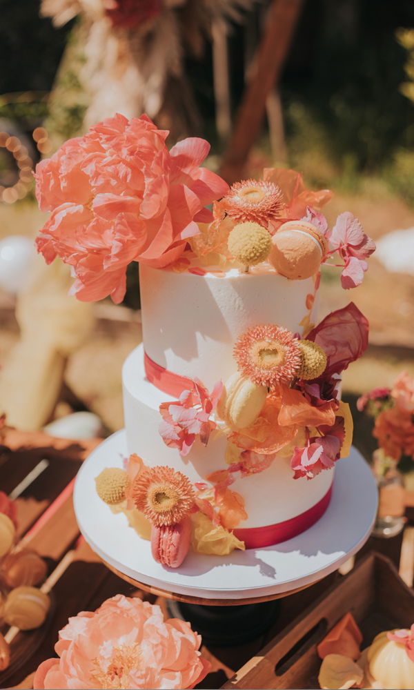 Lebhafte Hochzeitstorte mit zwei schlichten, weißen Ebenen, dekoriert mit üppigen Sommerblumen in warmen Farben. Die Akzente aus Pfingstrosen, Chrysanthemen, getrockneten Blättern und Macarons sorgen für eine verspielte, natürliche Optik. Rote Satinbänder unterstreichen das tropisch inspirierte Farbkonzept. Der rustikale Tisch und das Sonnenlicht verstärken das sommerliche Ambiente.