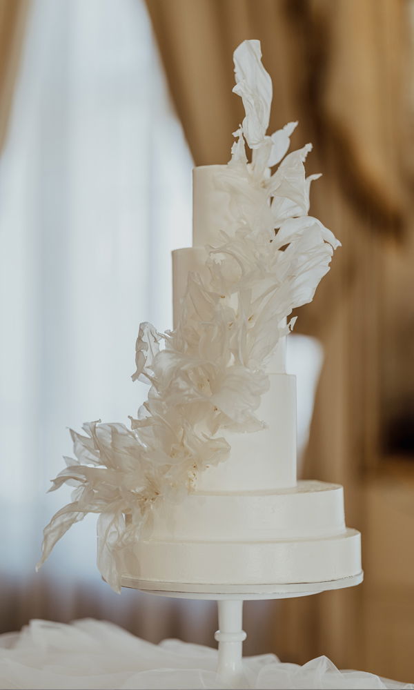Hochzeitstorte im Avantgarde-Stil mit vier runden Ebenen in glatter, weißer Optik. Die asymmetrisch angebrachten, hauchdünnen Reispapier-Dekorationen erinnern an schwebende Blütenblätter und verleihen der Torte eine künstlerische Note. Die elegante Umgebung unterstreicht den modernen Look.