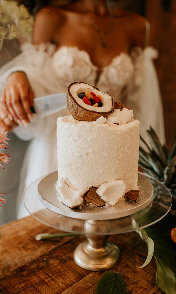 Eine exotische, einlagige Hochzeitstorte mit zarter Kokosflocken-Umhüllung und einer eindrucksvollen Kokosnuss-Dekoration auf der Spitze, gefüllt mit frischen Beeren. Die Szene wird von warmem, goldenem Licht durchflutet, während eine Braut in einem voluminösen Kleid gerade das Messer zum Anschneiden ansetzt.