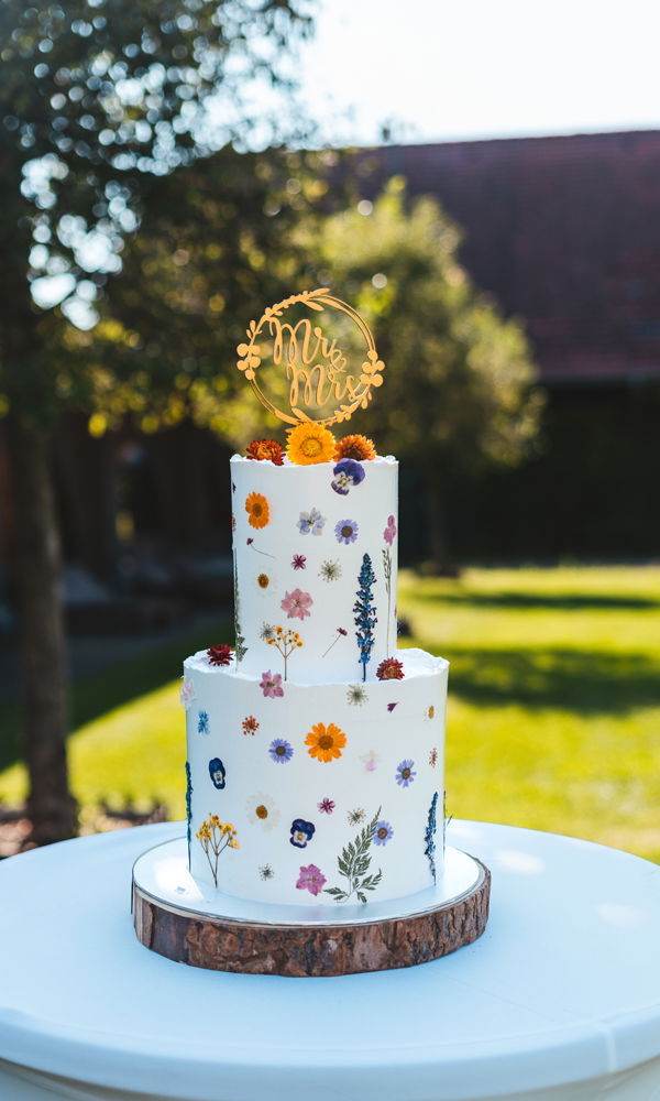 Elegante Hochzeitstorte im Vintage-Stil mit echten, gepressten Wildblumen auf zwei weißen Fondant-Etagen. Die florale Dekoration in kräftigen und pastelligen Farbtönen verleiht der Torte eine romantische, naturverbundene Ästhetik. Ein goldener Schriftzug „Mr & Mrs“ thront auf der oberen Etage, während der rustikale Holzsockel den natürlichen Look unterstreicht. Die Torte steht auf einem Tisch im Grünen mit weichem Sonnenlicht im Hintergrund.