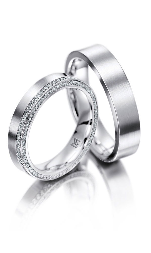 Moderne Hochzeitsringe aus Platin mit kühler, gebürsteter Oberfläche. Der Damenring wird von einer doppelten Reihe funkelnder Diamanten umrahmt, während das Herrenmodell eine puristische Eleganz ausstrahlt.