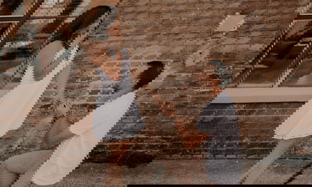 Romantischer Heiratsantrag vor einer Backsteinwand: Ein Mann kniet und präsentiert einen Verlobungsring, während die Frau emotional berührt ist.