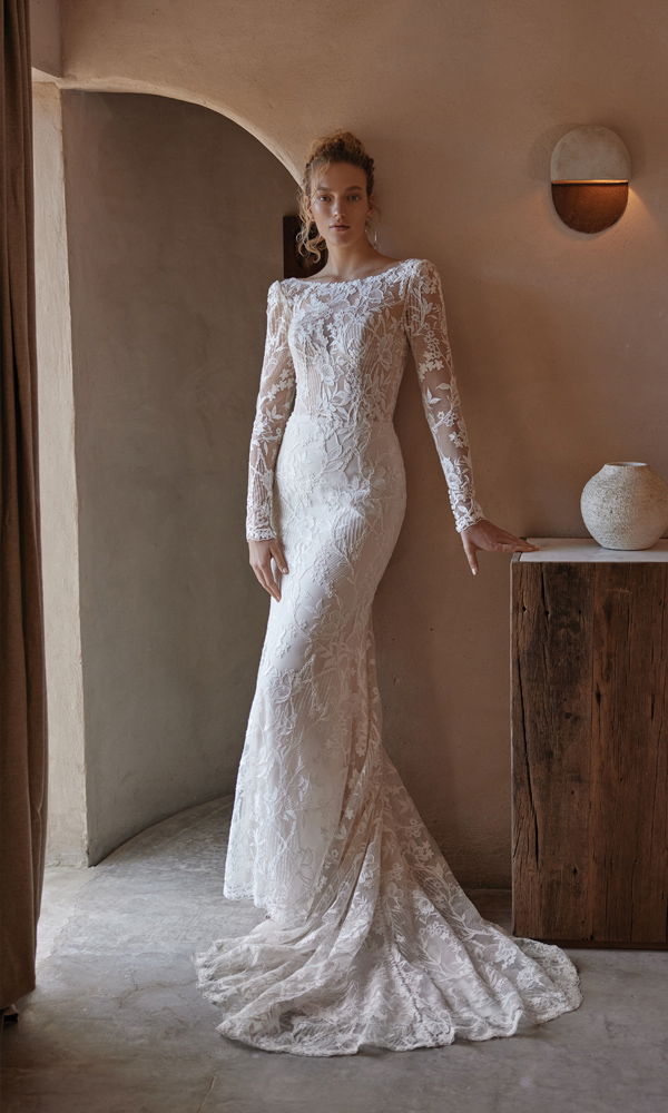 Feminines Brautkleid mit aufwendigen Spitzenverzierungen und eng anliegender Silhouette, aufgenommen vor einer natürlichen, warmen Wandkulisse.