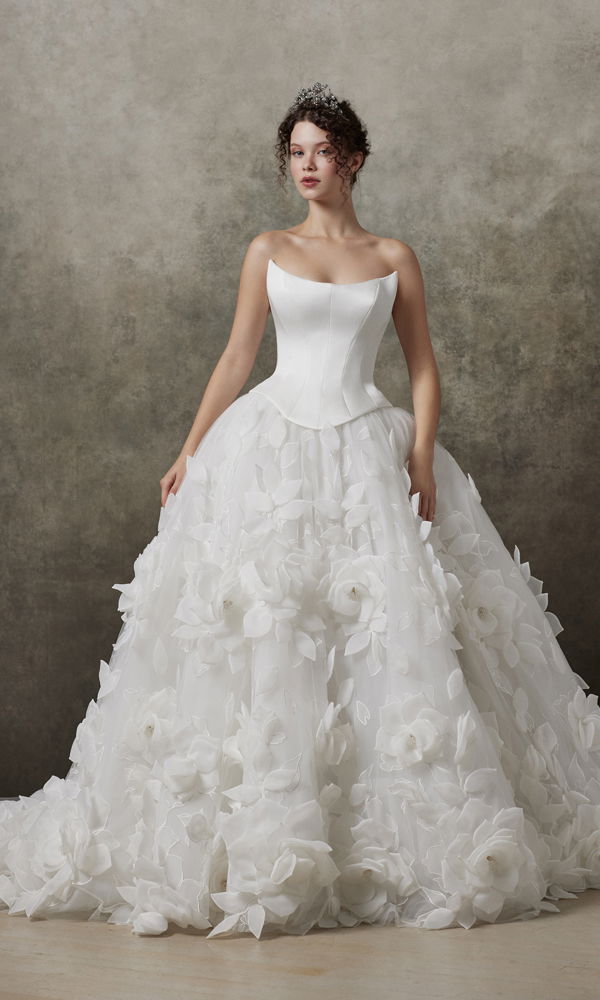 Romantisches Hochzeitskleid mit voluminösem Rock und kunstvollen Blütendetails, präsentiert vor einer klassischen, texturierten Kulisse.