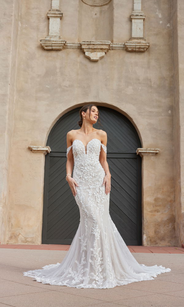 Hochzeitsporträt einer Frau in einem romantischen, schulterfreien Brautkleid mit detaillierter Blumenstickerei, inszeniert vor einem antiken Tor.