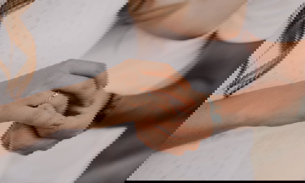 Detailaufnahme eines goldenen Verlobungsrings, getragen an der Hand einer Frau, zärtlich von ihrem Partner umfasst.