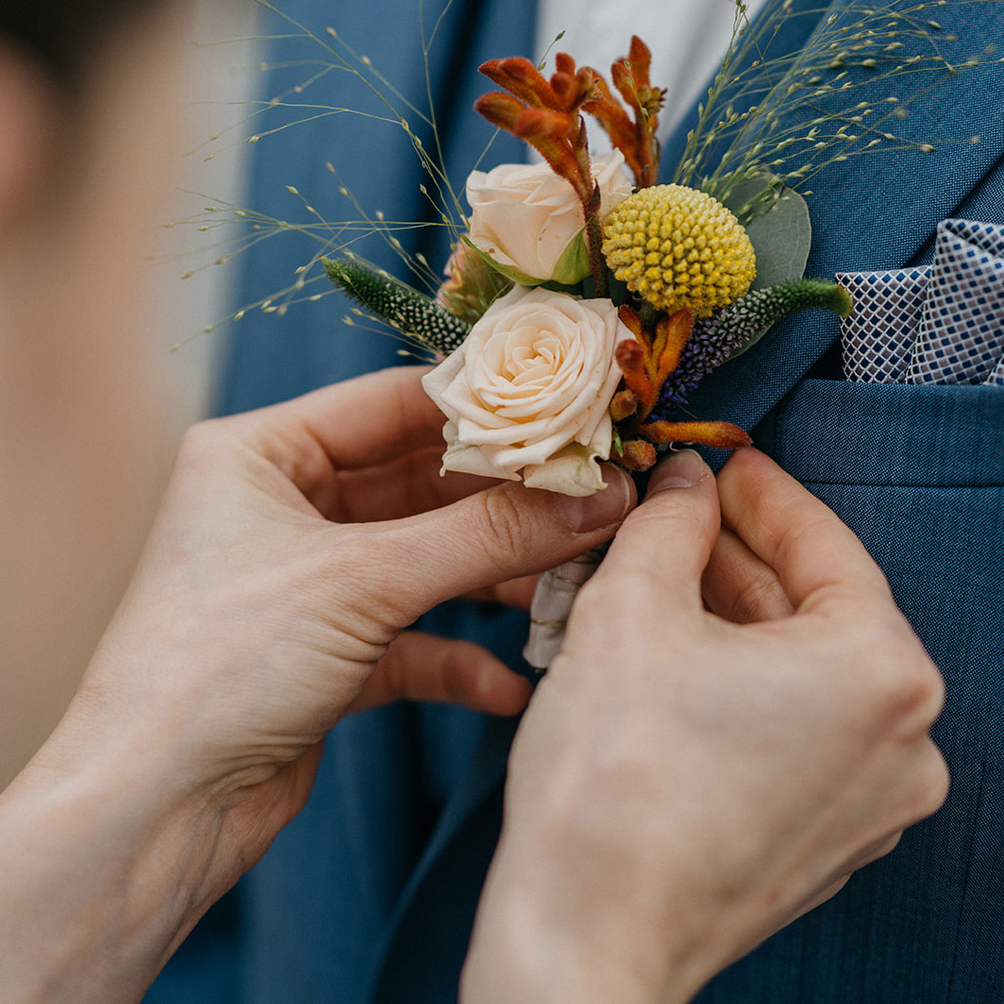 Hände befestigen eine florale Boutonniere an einem blauen Anzug. Die Blumenarrangements bestehen aus hellen Rosen, gelben und orangenen Elementen sowie feinen Gräsern und Details.