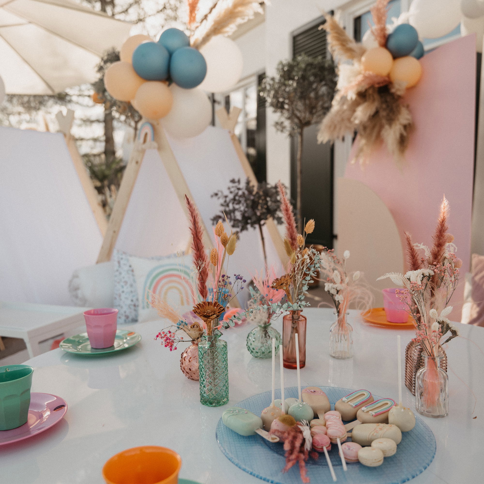 Detailreiche Party-Dekoration mit buntem Geschirr, Trockenblumen und pastellfarbenen Ballons, arrangiert auf einem Tisch unter freiem Himmel.