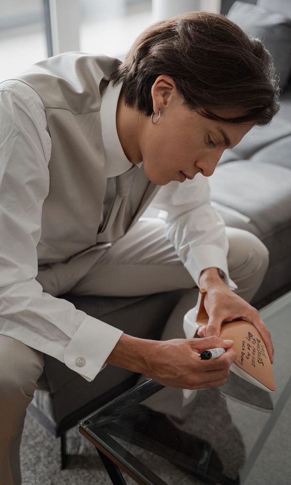 Ein Mann in einem hellen Anzug personalisiert einen Hochzeitsschuh mit einer handgeschriebenen Nachricht. Die Szene spielt sich auf einem eleganten Sofa ab.