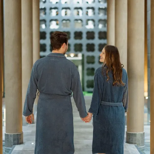 Paar in Bademänteln hält Händchen und geht durch einen stilvollen Spa-Gang mit Säulen und warmem Licht